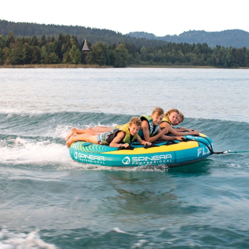 Three children on a blue and yellow Flight 4 boat tube in a lake with trees and mountains in the background.