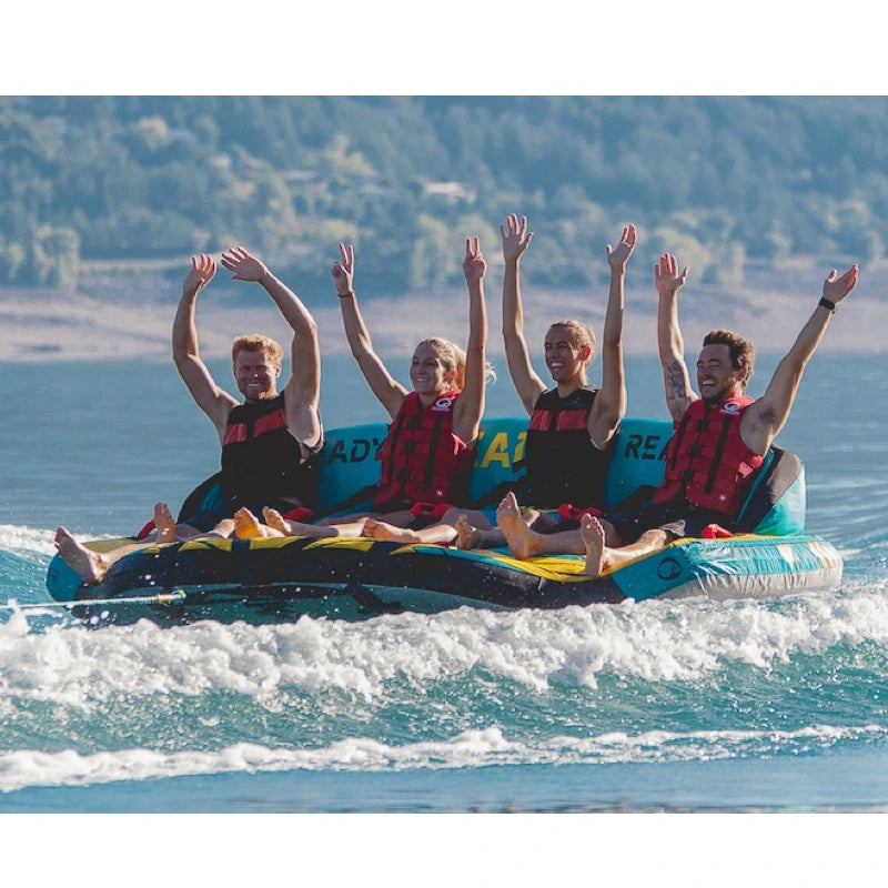 Four people on a boat tube with raised arms in the water, surrounded by trees and mountains.