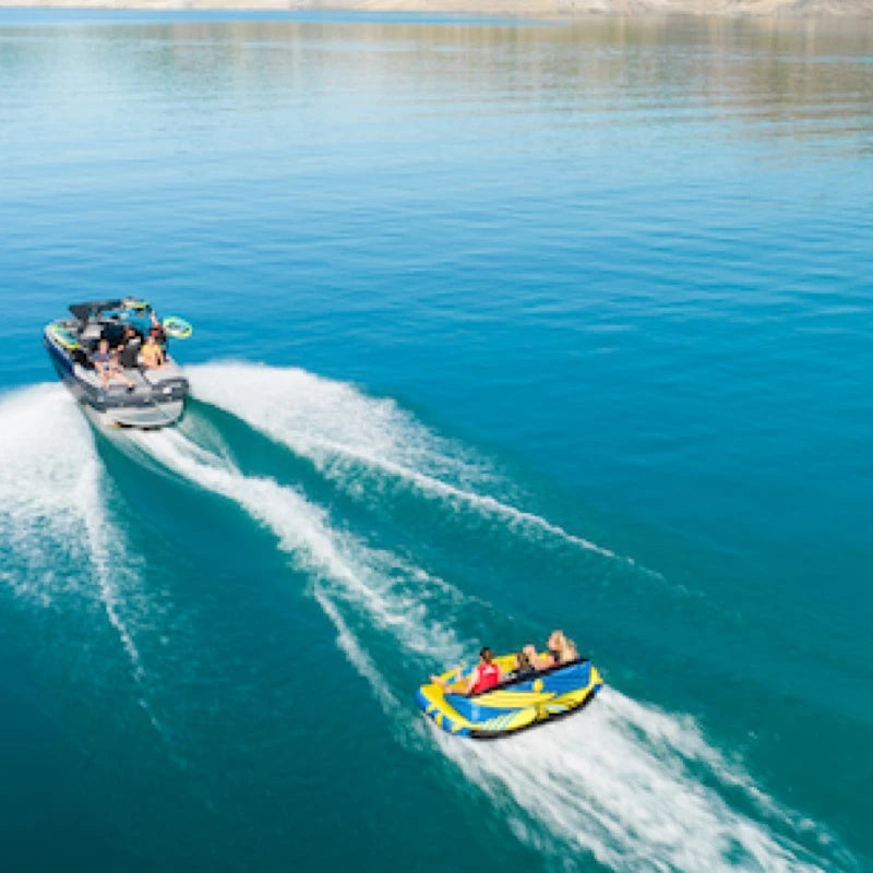 Speedboat towing a yellow and blue inflatable tube with people on a clear blue lake.