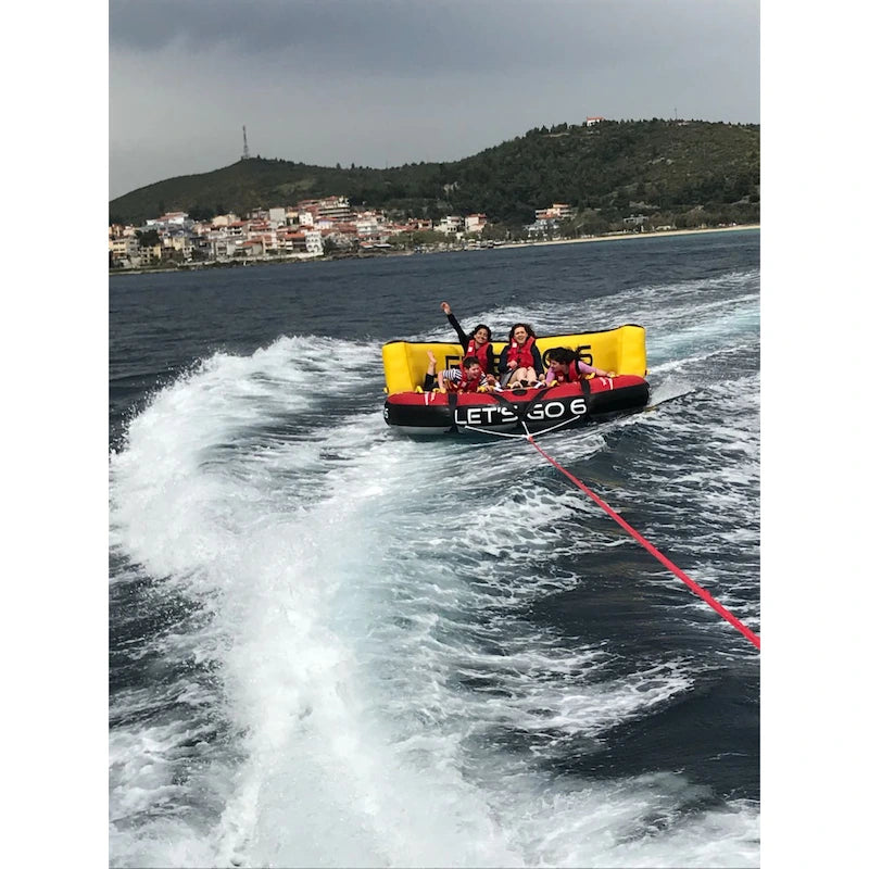 People on an inflatable tube being pulled by a boat on a body of water with a town in the background.