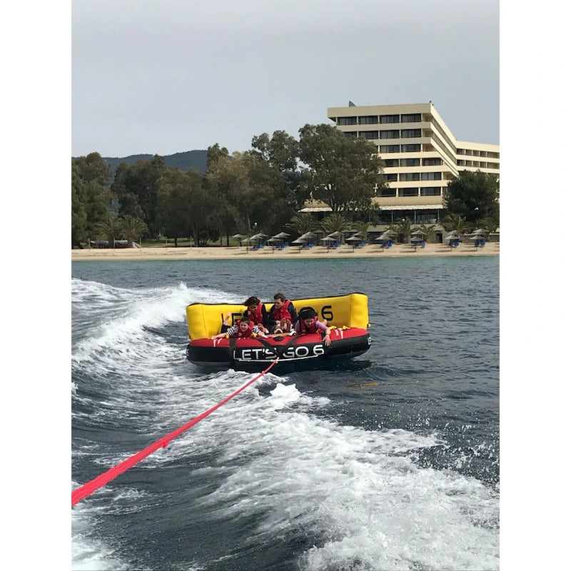 Inflatable water tube with people being pulled by a boat on a body of water with a building and trees in the background.