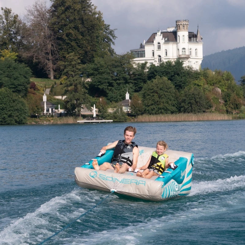 Two people on a water tube in a lake with a castle-like building in the background