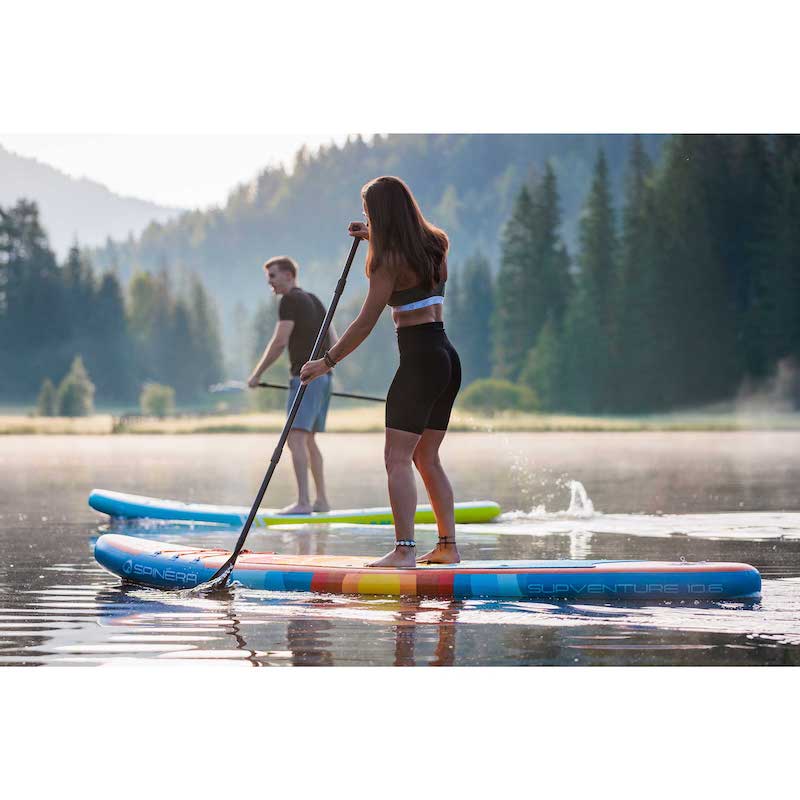 Two people paddleboarding on a lake with a scenic background of trees and mountains.
