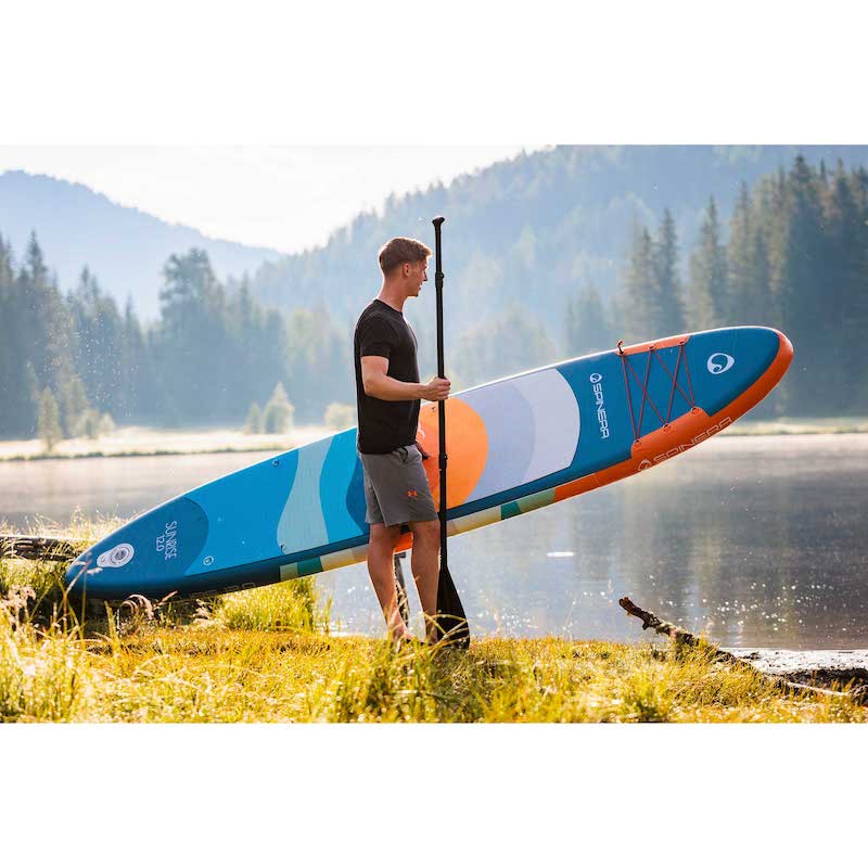 Man holding a colorful Spinera Supventure sunrise paddleboard on a grassy area with a lake and mountains in the background