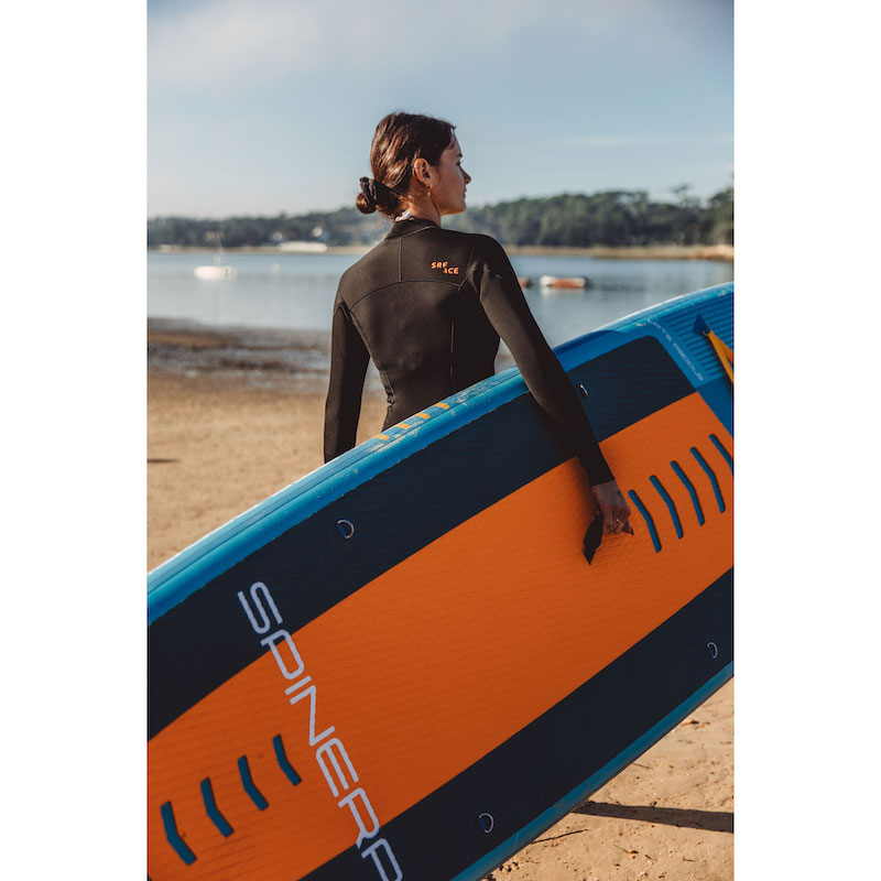 Person carrying an orange and blue paddleboard with 'SPINERA' branding on a beach.