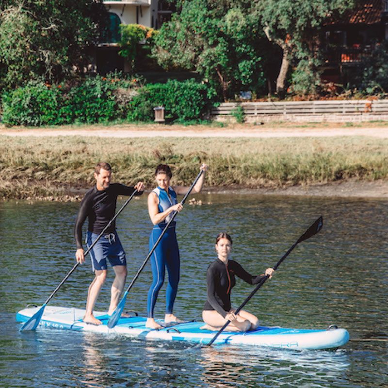 Three people paddleboarding on a lake with trees and a path in the background.