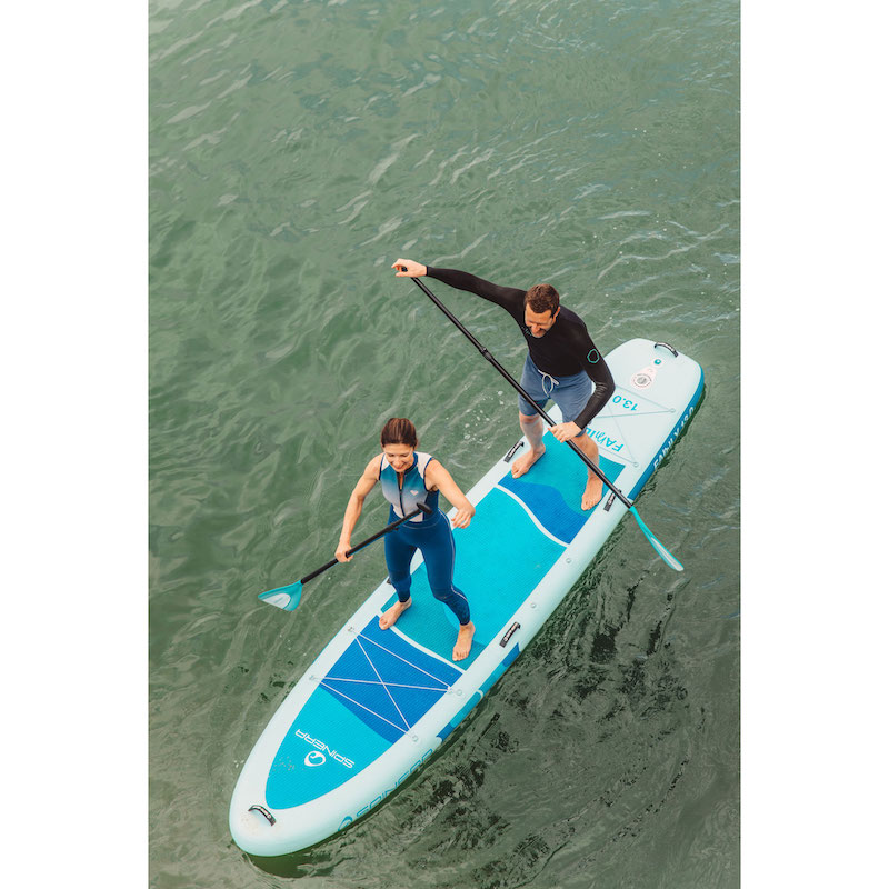 Two people paddleboarding on a blue and white inflatable stand-up paddleboard in green water.