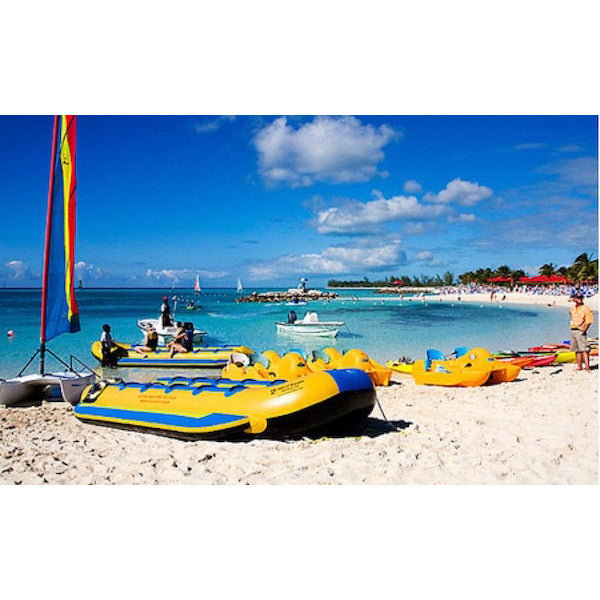 Side view of the yellow and blue Island Hopper 12 Person Towable Banana Boat Taxi sitting on the beach next to the ocean. 