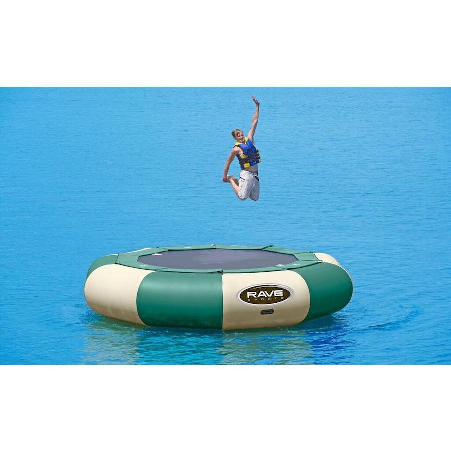A guy jumping on a green and tan Rave Aqua Jump Eclipse 150 Northwoods Water Trampoline on the lake. 