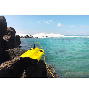 Chasing Gladius Mini Underwater Drone sits on a large rock next to the ocean as waves crash in the background.  You can see the details of the yellow underwater drone for sale and the yellow tether coming out of the black connector to the underwater drones for sale.