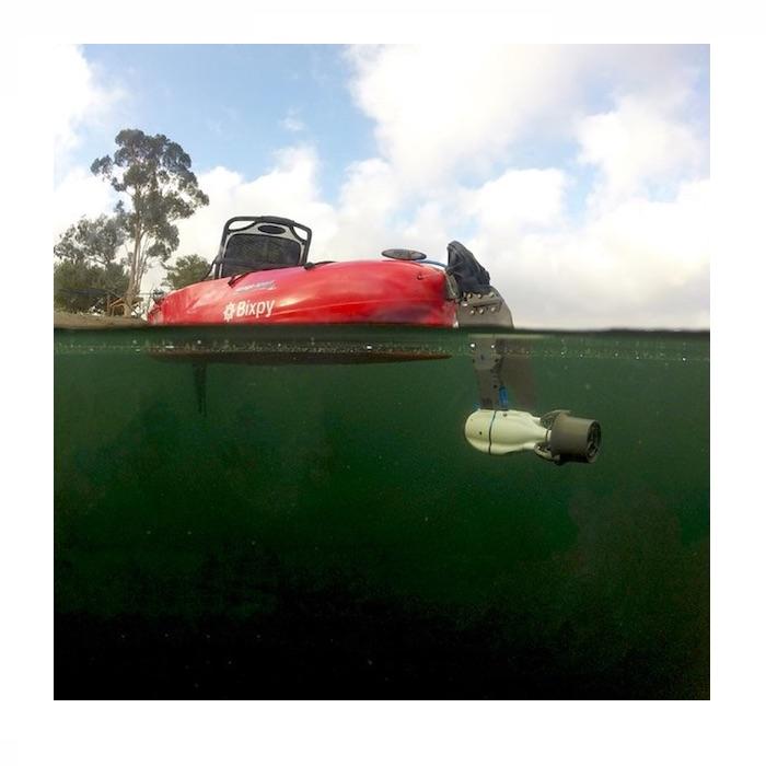 Bixpy Hobie Twist & Stow Kayak Rudder Adapter shown attached to the back of a red Hobie kayak floating in a lake.  Split shot camera angle shows the black Hobie Twist & Stow attached to the grey and black Jet Motor underwater. 