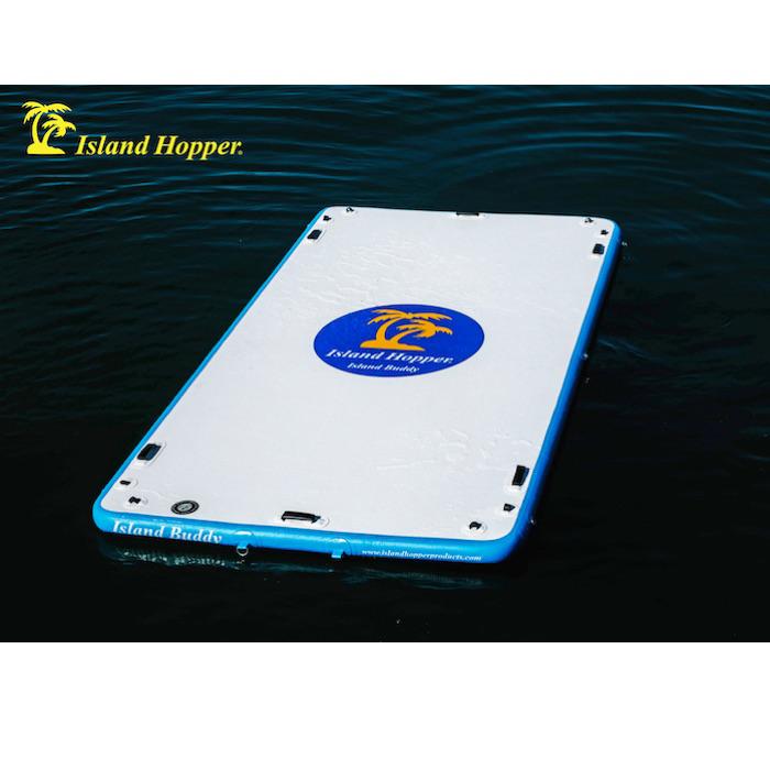 Island Hopper 12ft Island Buddy Inflatable Water Mat sitting in the middle of the lake by itself. It is white on top with a royal blue oval. Inside the oval is yellow palm trees and white Island Hopper Island Buddy lettering in the oval and on the sides.