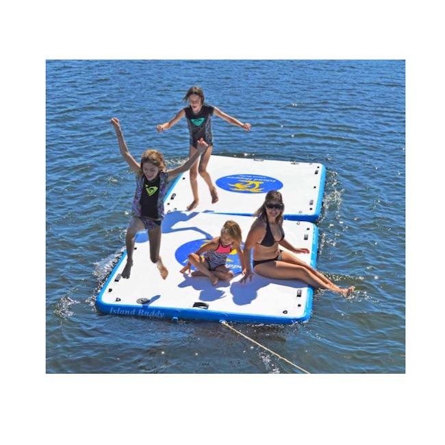 Two Island Hopper Island Buddy Floating Swim Platform hooked together in the water and 2 young kids are running and jumping off of the end.  Another small childs sits next to mom watching the others run and jump off of the floating swim platform that is white on top with blue Island Hopper Island Buddy logo with yellow lettering.