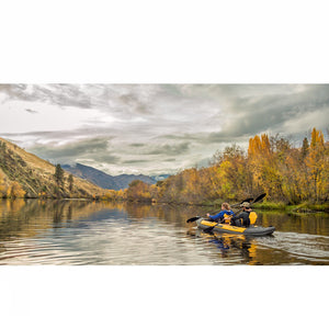 Man and woman paddling the yellow and grey Advanced Elements Island Voyage 2 Tandem Inflatable Kayak down a river in a picturesque scene. 