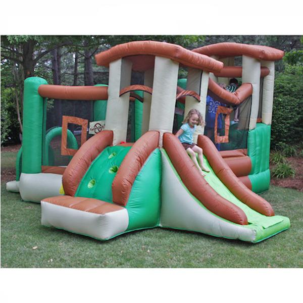 KidWise Clubhouse Climber Bounce House side view showing 2 slides, with 1 girl sliding down 1 slide.  Catwalk and ball pit in the background, bounce house against a forest.