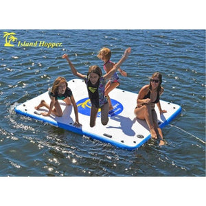 Island Hopper Island Buddy Inflatable Floating swim platform is being used by 4 kids jumping off of it into the lake.  The inflatable water platform has a white top with blue circle in the middle.  The Island Hopper floating swim platform is floating attached only by a rope, water surrounds the floating swim platform.