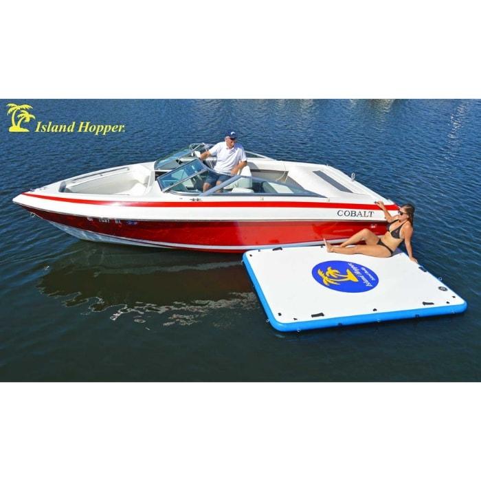 Island Hopper Island Buddy Inflatable Floating swim platform is tied up to a boat while a woman kicks back and relaxes.  The red boat is sitting idle in the water while the white and blue floating swim platform rests against it.