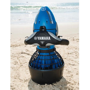 A Yamaha RDS250 Seascooter sits upright in the sand at the beach with the ocean visible in the background.  Yamaha is clearly printed in white letters across the handle bars.  There are also grey highlights on the body and handles.