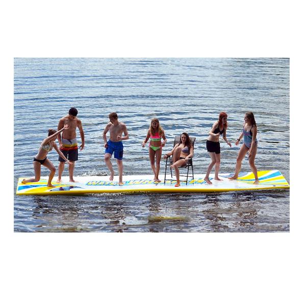 Rave Water Whoosh 15 Floating Water Mat on the lake with 6 people standing and playing and one girl sitting in a chair on the sturdy inflatable water mat.  The floating swim mat is white and yellow with blue highlights.