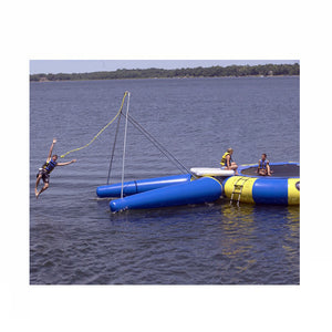 Man letting go over the rope on a Blue Rave Rope Swing Water Trampoline Attachment attached to a blue and yellow Rave Water Trampoline.  Rope Swing water trampoline attachment is on a dark lake against a blue sky. 