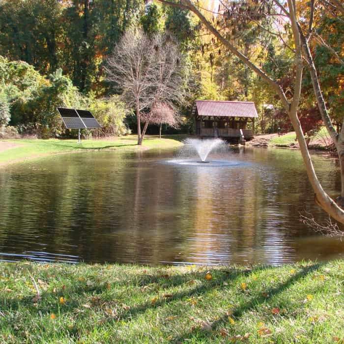 Scott Aerator DA-20 Display Floating Solar Pond Aerator 3/4 Hp Floating Aerator Fountain.  This small pond aerator fountain is in a small farm pond.  Blue/brown water wit a covered bridge at the end of the pond. The black floating solar pond aerator power panel is on the left side of the lake. 