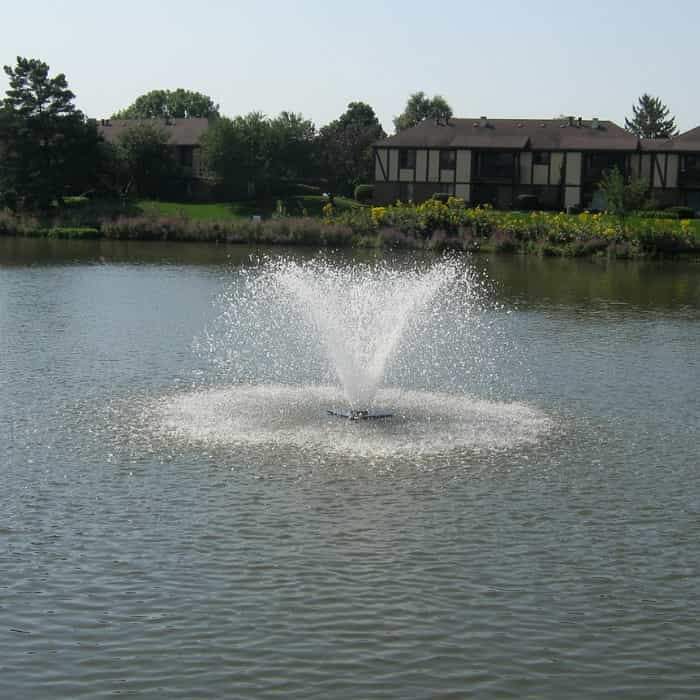Scott Aerator DA-20 Display Floating Solar Pond Aerator 3/4 Hp Floating Aerator Fountain.  This small pond aerator fountain is in a small farm pond.  Blue/brown water wit a covered bridge at the end of the pond. The black floating solar pond aerator power panel is on the left side of the lake. 