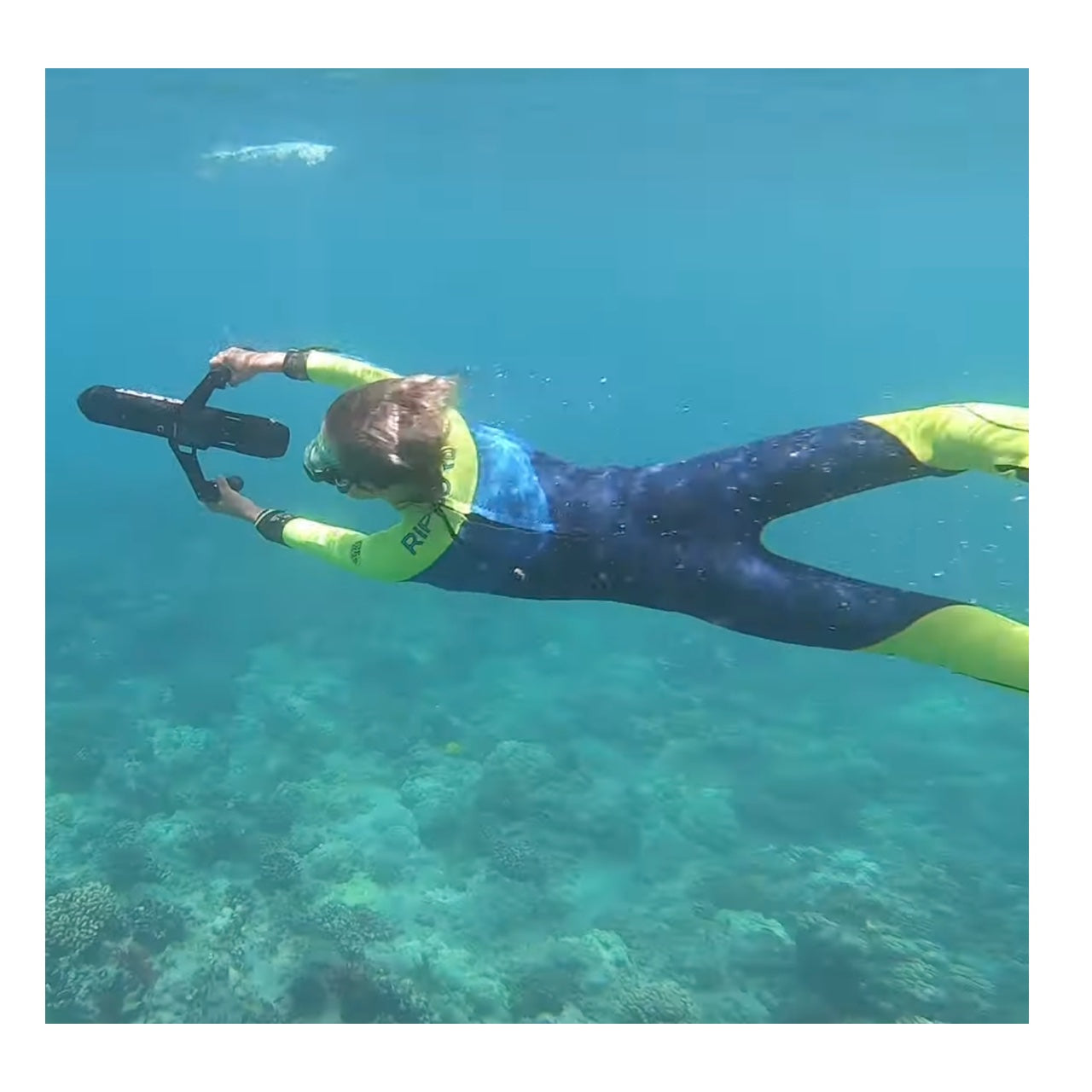 A young girl uses the ScubaJet Pro with Underwater Kit so she can use the underwater scooter to explore the ocean. She is wearing a bright wetsuit and we see their view from the top.