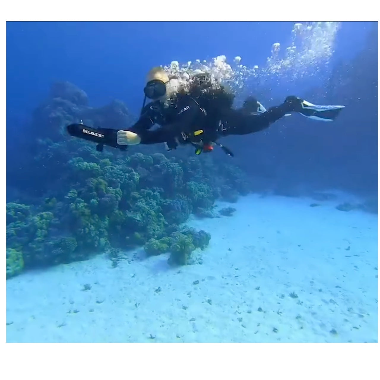 A diver uses a ScubaJet pro near the floor of the ocean. He holds the all black with silver lettering ScubaJet Pro in both hands and air bubles come out of his mask and regulator.