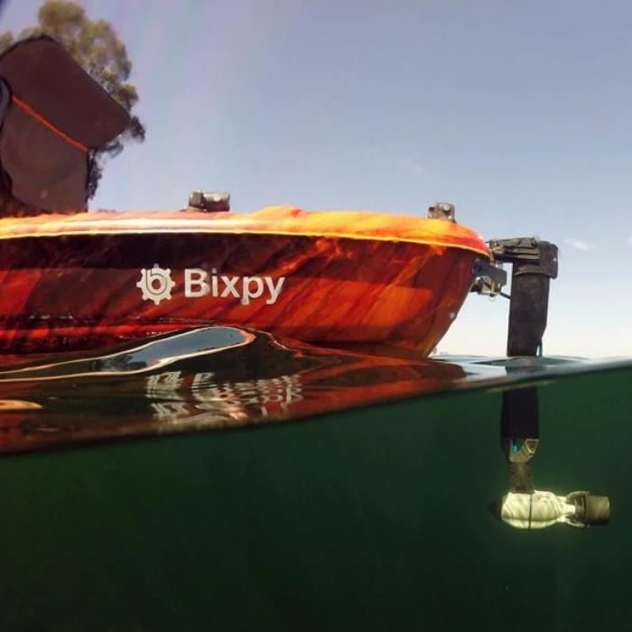 Bixpy Kayak Jet Motor for sale is shown in use on an orange Bixpy Kayak.  The Bixpy Jet Thruster is attached to the rear end of the kayak using the Bixpy Universal Rudder Adapter.  You can see the grey, black, and blue Bixpy Thruster through the water attached to the black Bixpy Universal Rudder Adapter.  It is shown here trolling and not creating a noticeable thrust in the water.  This is one of the best trolling motor for kayak that you will find.