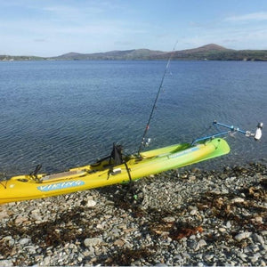 Bixpy Kayak Jet Motor is shown in use with a Bixpy Universal Kayak and Canoe Adapter on a green and yellow kayak.  The Bixpy Jet Thruster is fully out of the water and the kayak is resting on the rocky shore on a stand.  The Kayak Outboard Motor Power Pack is not visible, but the blue connecting cable is.