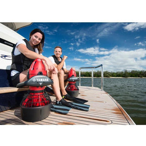 A couple sits on the back of a boat on a wood swim deck each with their red Yamaha RDS200 Sea Scooter in hand.  