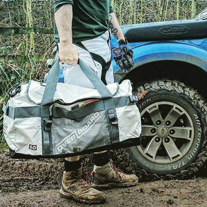 This is an image of the Adventure Weatherproof Duffel Bag in action being held by a man standing on a muddy environment.